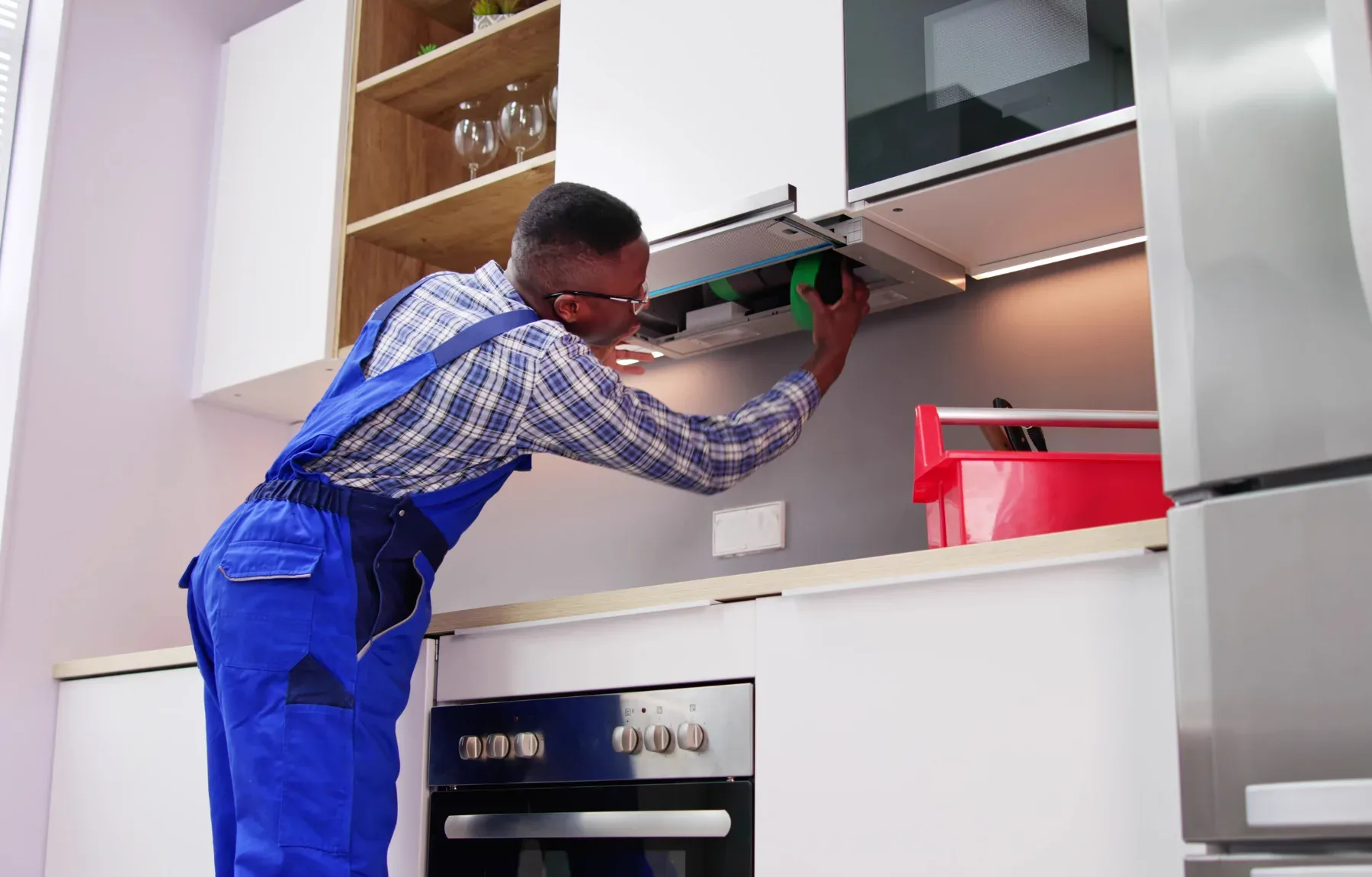 A man in blue coveralls and a plaid shirt, wearing glasses, reaches up to install or repair a range hood built into white kit