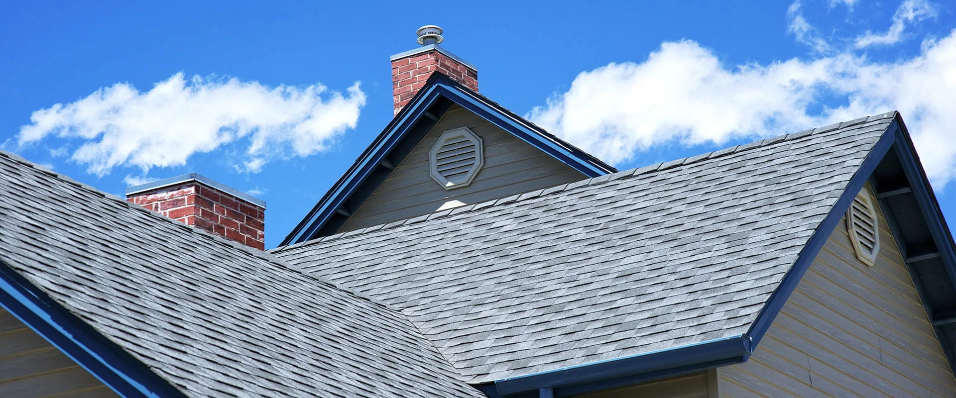 Residential roof with gray shingles, brick chimneys, and blue trim against clear sky.
