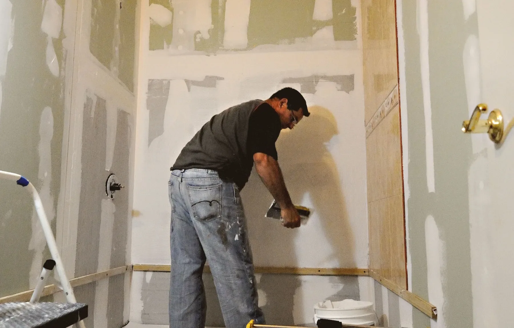 A worker in a dark gray t-shirt and blue jeans bends over to apply white joint compound to a wall with a putty knife, smoothi