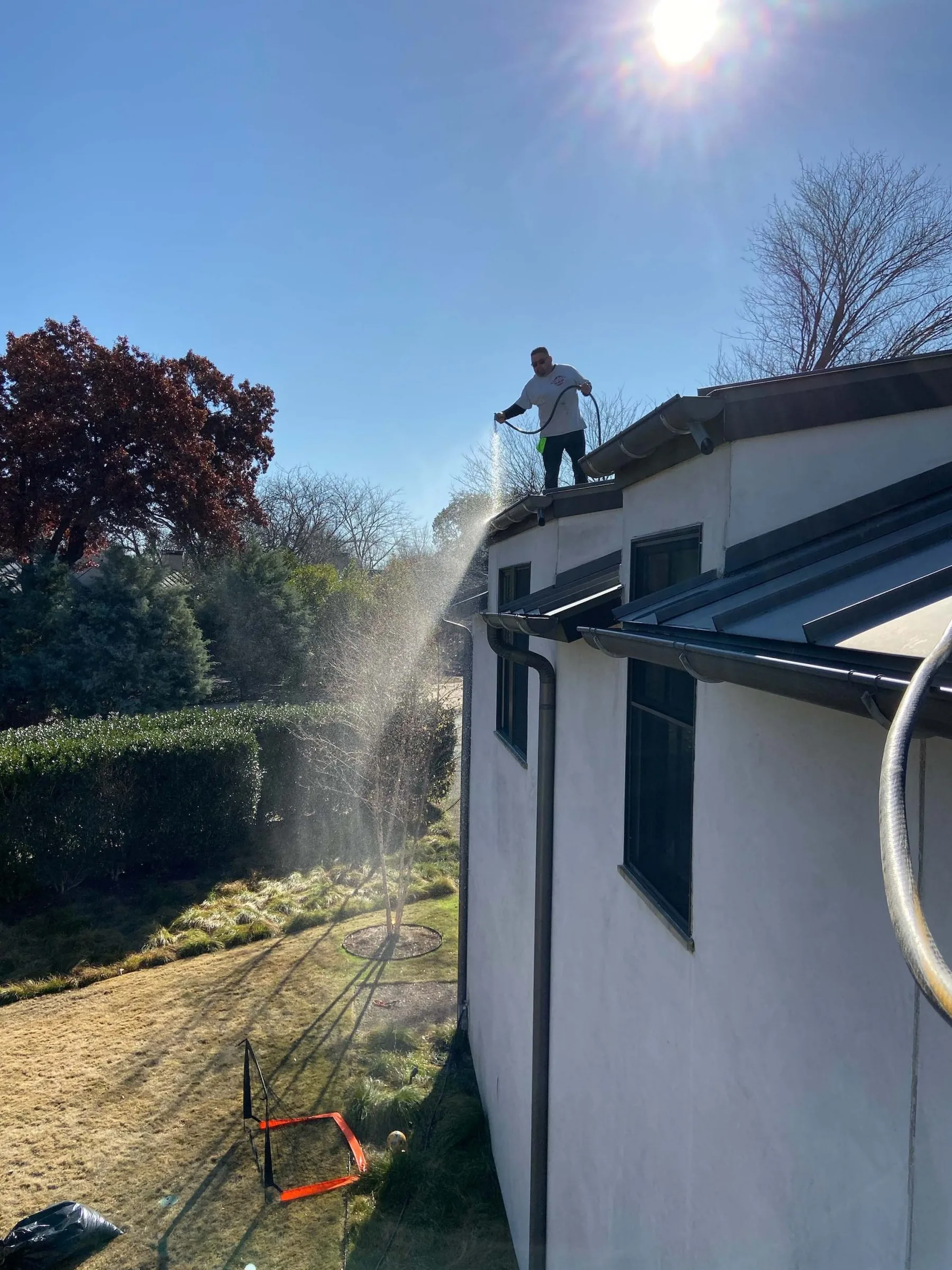 Worker on residential roof cleaning gutters with pressure washer spray on sunny day.