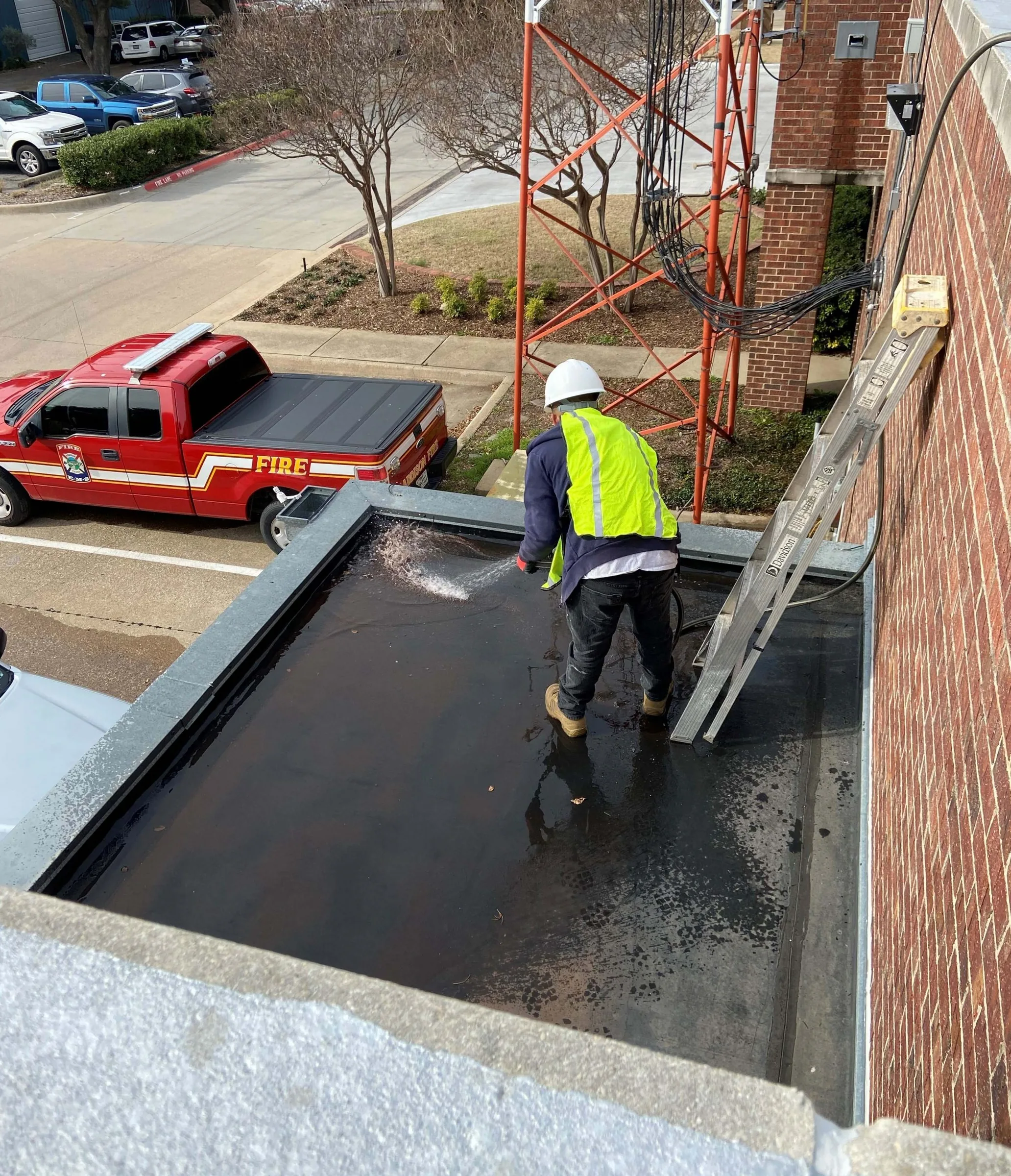 Worker in safety gear cleaning a flat roof gutter next to a brick building.